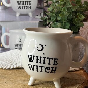 May include: Cream ceramic mug with a rounded body, handle, and three small feet. The mug has the text "WHITE WITCH" in black, with a crescent moon and stars design. The mug is displayed on a wooden surface.
