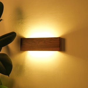 Puede incluir: Aplique de pared rectangular de madera con una luz cálida que emana del centro. El aplique está montado en una pared de color claro, con una planta en primer plano. La madera tiene una veta natural.