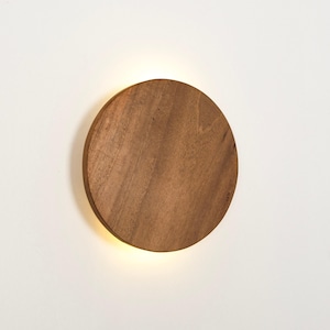 Puede incluir: Aplique de pared redondo de madera con veta natural. La luz emite un brillo suave desde el borde inferior. El diseño es minimalista y moderno, adecuado para varios estilos de interior. El diámetro es de aproximadamente 20 cm.