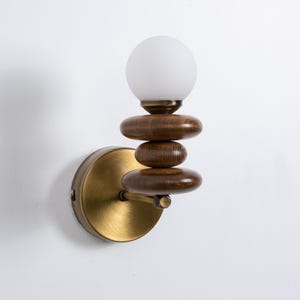 May include: A brass wall sconce with a frosted glass globe and a stack of four wooden discs. The discs are stacked in a staggered pattern, creating a unique and modern design.