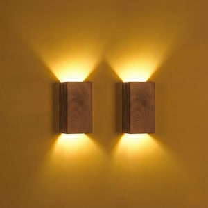 May include: Two wooden wall sconces with a warm, inviting glow. The sconces are rectangular, with a natural wood grain finish. The light shines both up and down, creating a cozy ambiance. The wall is a warm yellow color.