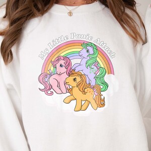 May include: White sweatshirt with a rainbow and three My Little Pony figures. The text on the sweatshirt reads "My Little Panic Attack".