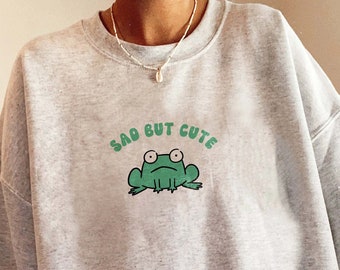 May include: A gray sweatshirt with a green frog graphic and the text "Sad But Cute".