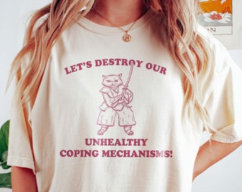 May include: A cream coloured t-shirt with a pink graphic of a cat holding a sword. The text on the shirt reads "Let's destroy our unhealthy coping mechanisms!"