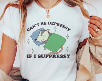 May include: White t-shirt with a graphic of a possum sleeping in a blanket with the text "Can't be Depressy if I Suppressy".