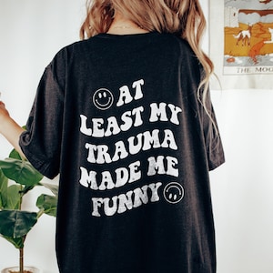 Op de afbeelding: Zwart oversized T-shirt met een witte grafische afbeelding met de tekst "At Least My Trauma Made Me Funny" met een smiley aan beide kanten van de tekst.