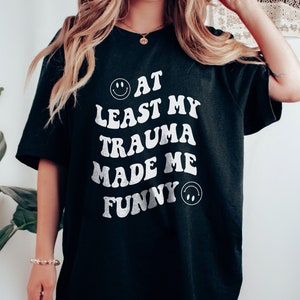May include: Black oversized t-shirt with white text that reads "At Least My Trauma Made Me Funny" with a smiley face on either side of the text.