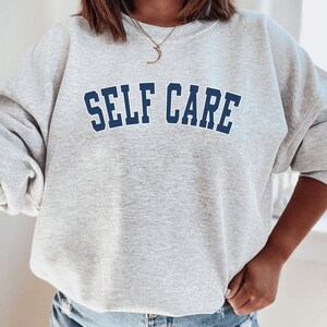 Peut inclure: Sweat-shirt gris avec le texte "SELF CARE" en lettres bleues soulign&eacute;es.