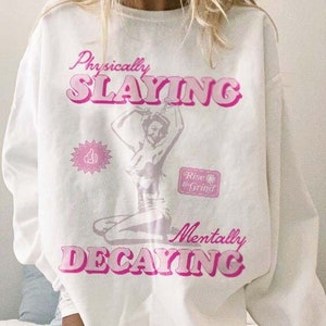 May include: White long-sleeve sweatshirt with a pink graphic print that says "Physically Slayin'" and "Mentally Decaying". The graphic also includes a drawing of a woman with a thumbs up and the text "Rise & Grind".