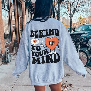 K&ouml;nnte beinhalten: Grauer Pullover mit einem Grafikdruck eines Herzens mit Augen und Beinen. Der Text auf dem Pullover lautet "Be Kind To Your Mind".