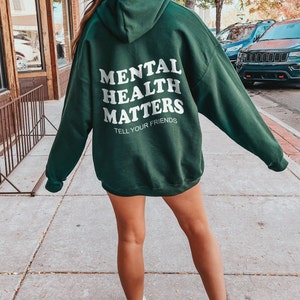 K&ouml;nnte beinhalten: Ein dunkelgr&uuml;ner Kapuzenpullover mit dem Text "Mental Health Matters" und "Tell Your Friends" in wei&szlig;en Buchstaben auf der R&uuml;ckseite.