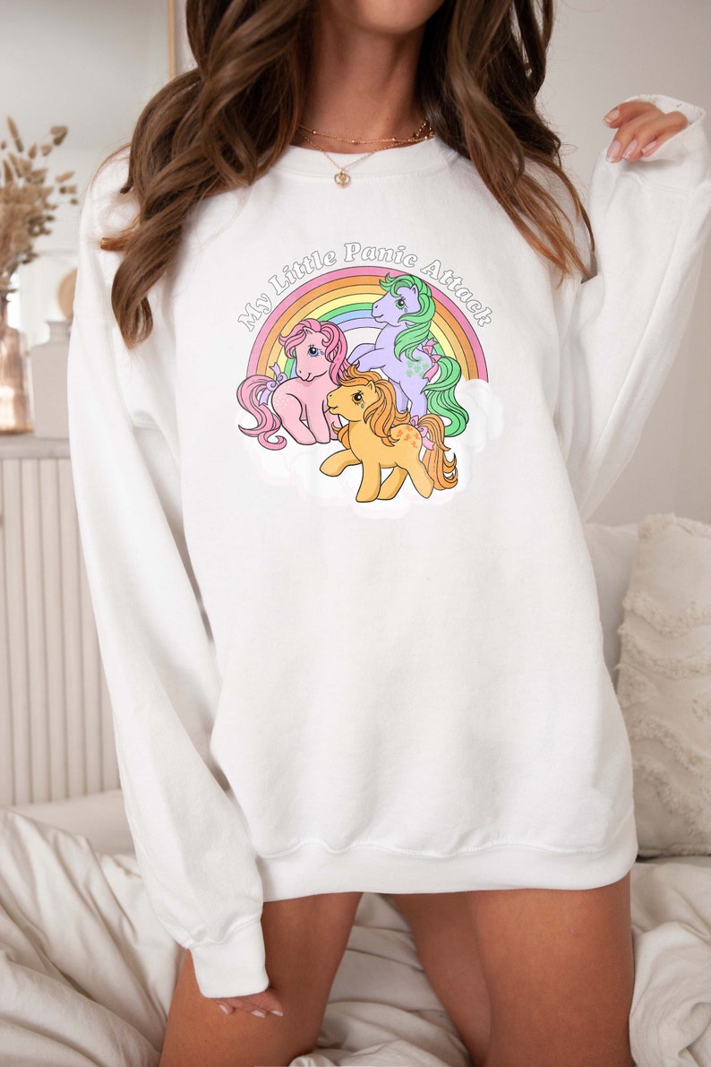 May include: White sweatshirt with a rainbow and three My Little Pony figures. The text on the sweatshirt reads "My Little Panic Attack".
