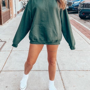 K&ouml;nnte beinhalten: Ein &uuml;bergro&szlig;er, dunkelgr&uuml;ner Hoodie mit einer Kapuze mit Kordelzug und einer K&auml;ngurutasche. Der Hoodie wird von einer Person mit wei&szlig;en Turnschuhen getragen.