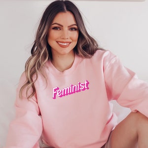 K&ouml;nnte beinhalten: Ein rosa Sweatshirt mit dem Wort "Feminist" in rosa und wei&szlig;en Buchstaben.
