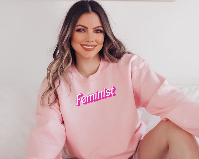 K&ouml;nnte beinhalten: Ein rosa Sweatshirt mit dem Wort "Feminist" in rosa und wei&szlig;en Buchstaben.