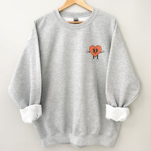 K&ouml;nnte beinhalten: Ein grauer Pullover mit einem roten Herz-Motiv auf der Vorderseite. Das Herz hat ein Gesicht mit Augen, einem L&auml;cheln und zwei Daumen nach oben.
