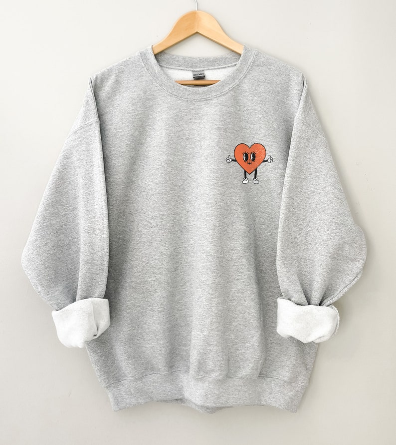 K&ouml;nnte beinhalten: Ein grauer Pullover mit einem roten Herz-Motiv auf der Vorderseite. Das Herz hat ein Gesicht mit Augen, einem L&auml;cheln und zwei Daumen nach oben.