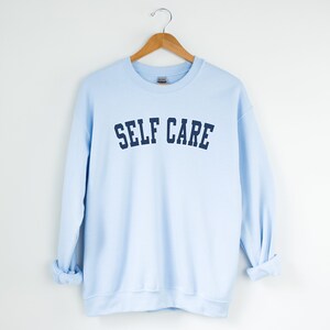 Peut inclure: Un sweat-shirt bleu clair avec le texte "SELF CARE" imprim&eacute; en bleu marine.