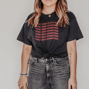 May include: A black t-shirt with the phrase "You are enough" repeated in red lettering. The shirt is tied at the waist and worn with dark wash jeans.