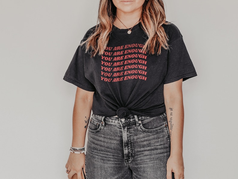 May include: A black t-shirt with the phrase "You are enough" repeated in red lettering. The shirt is tied at the waist and worn with dark wash jeans.