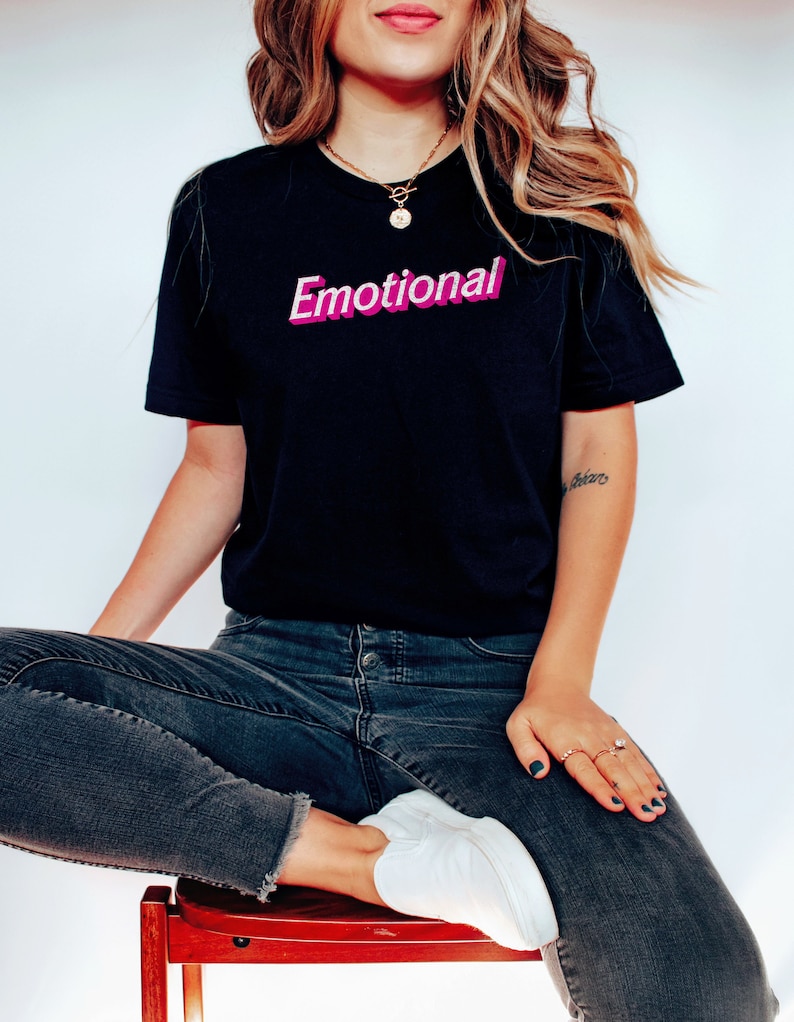 May include: A woman wearing a black t-shirt with the word "Emotional" printed in pink and white letters. She is wearing blue jeans and white sneakers.