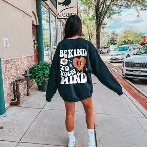K&ouml;nnte beinhalten: Ein schwarzer Pullover mit einem wei&szlig;en Grafikdruck, der "Be Kind to Your Mind" mit einem Comic-Herz und einer darauf zeigenden Hand sagt. Die Person, die den Pullover tr&auml;gt, geht auf einem B&uuml;rgersteig vor einem Geb&auml;ude entlang.