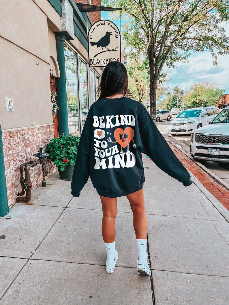K&ouml;nnte beinhalten: Ein schwarzer Pullover mit einem wei&szlig;en Grafikdruck, der "Be Kind to Your Mind" mit einem Comic-Herz und einer darauf zeigenden Hand sagt. Die Person, die den Pullover tr&auml;gt, geht auf einem B&uuml;rgersteig vor einem Geb&auml;ude entlang.