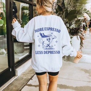 Puede incluir: Sudadera gris con un gr&aacute;fico azul de una taza de caf&eacute; con una cara y el texto "More Espresso Less Depresso".