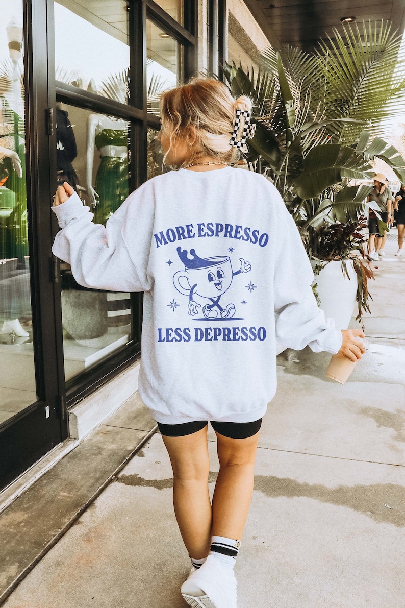 Puede incluir: Sudadera gris con un gr&aacute;fico azul de una taza de caf&eacute; con una cara y el texto "More Espresso Less Depresso".