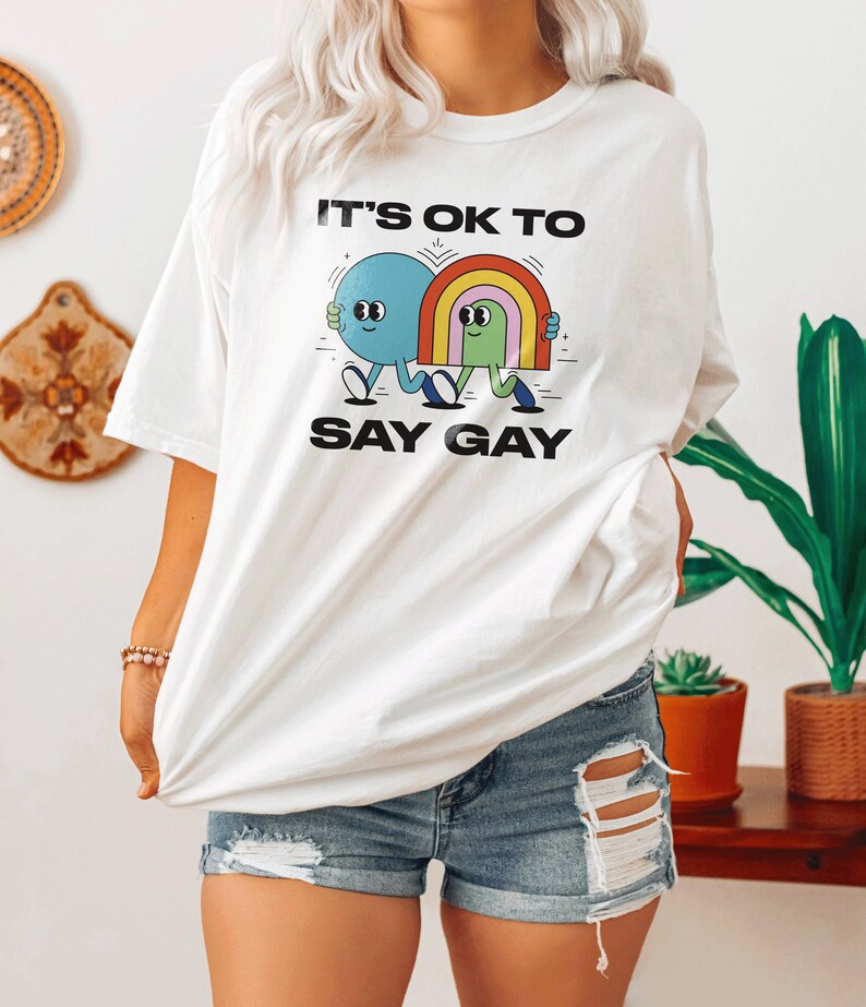 May include: White t-shirt with a graphic of two cartoon figures holding hands. The text "It's ok to say gay" is printed on the shirt.