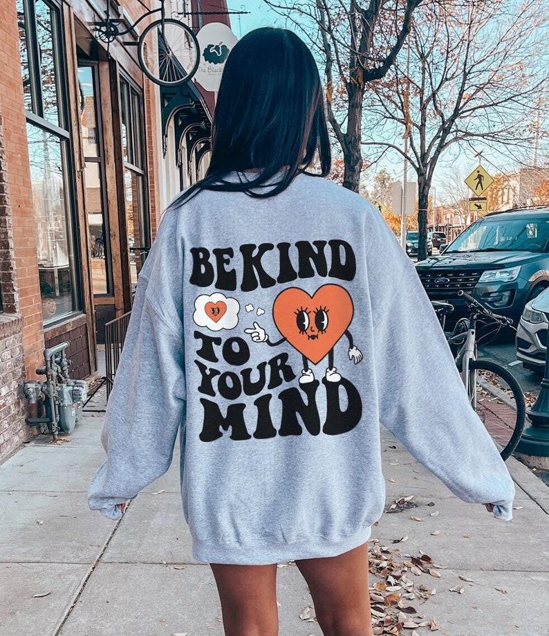 K&ouml;nnte beinhalten: Grauer Pullover mit einem Grafikdruck eines Herzens mit Augen und Beinen. Der Text auf dem Pullover lautet "Be Kind To Your Mind".