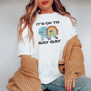 May include: White t-shirt with a graphic of two cartoon characters holding hands. The text "It's ok to say gay" is printed below the graphic.
