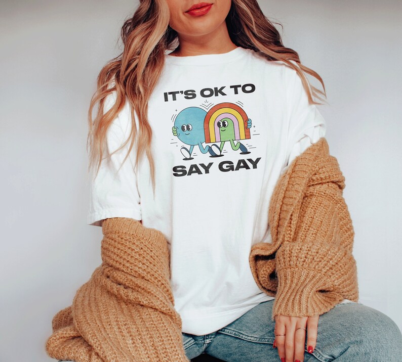 May include: White t-shirt with a graphic of two cartoon characters holding hands. The text "It's ok to say gay" is printed below the graphic.