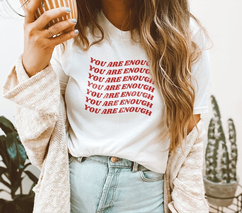 May include: White t-shirt with the text "YOU ARE ENOUGH" repeated in red letters in a retro font.