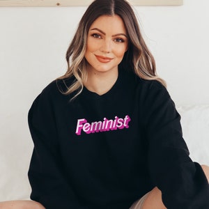 K&ouml;nnte beinhalten: Ein schwarzer Pullover mit einem pinkfarbenen "Feminist"-Aufdruck auf der Vorderseite.