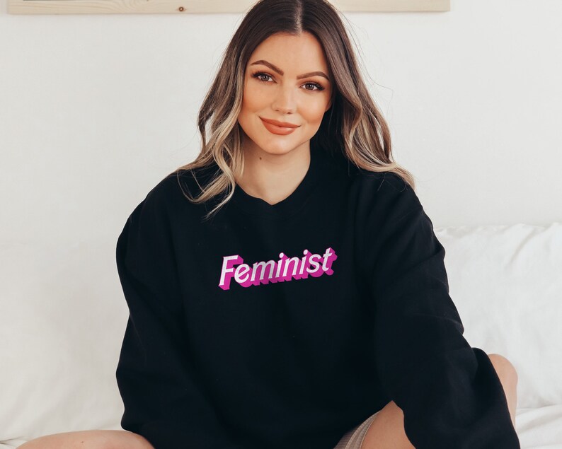 K&ouml;nnte beinhalten: Ein schwarzer Pullover mit einem pinkfarbenen "Feminist"-Aufdruck auf der Vorderseite.
