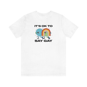 May include: White t-shirt with a graphic of two cartoon characters walking. The characters are holding hands and walking under a rainbow. The text "It's ok to say gay" is printed below the graphic.