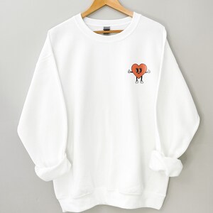 K&ouml;nnte beinhalten: Wei&szlig;er Pullover mit einem roten Herz-Motiv auf der linken Brust. Das Herz hat ein l&auml;chelndes Gesicht und zeigt mit beiden H&auml;nden nach oben.