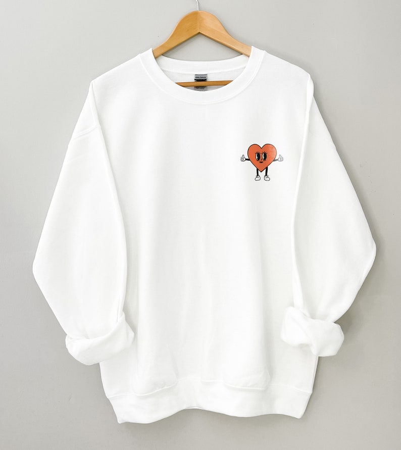 K&ouml;nnte beinhalten: Wei&szlig;er Pullover mit einem roten Herz-Motiv auf der linken Brust. Das Herz hat ein l&auml;chelndes Gesicht und zeigt mit beiden H&auml;nden nach oben.