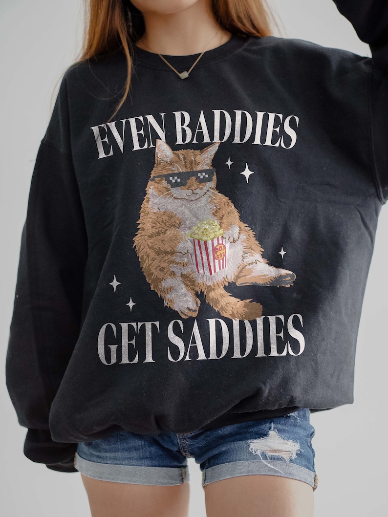 May include: Black sweatshirt with a white graphic of a cat wearing sunglasses and holding a bucket of popcorn. The text "EVEN BADDIES GET SADDIES" is printed on the sweatshirt.