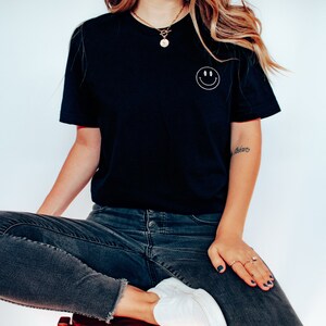 Op de afbeelding: Een vrouw draagt een zwart T-shirt met een witte smiley die op de borst is geborduurd. Ze draagt een donkerblauwe jeans en witte sneakers. Ze zit op een houten kruk.