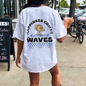 May include: White t-shirt with the text "Happiness Comes in Waves" and a cartoon sun surfing on a wave. The shirt also has the text "It Will Find You Again" and the phrases "Be Kind to Your Mind", "Mental Health Matters", and "Keep on Going".