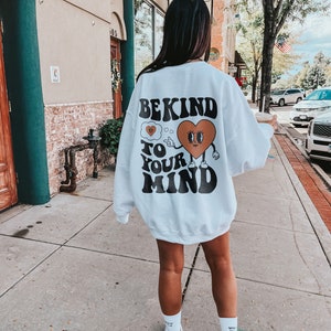 K&ouml;nnte beinhalten: Wei&szlig;er Pullover mit einem grafischen Aufdruck eines Comic-Herzens mit Augen und einer Gedankenblase, auf der "Be Kind To Your Mind" steht.