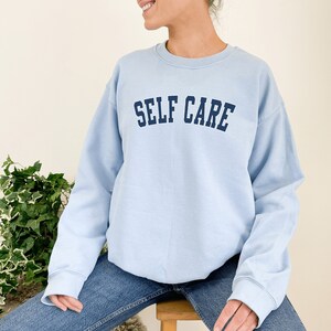 Peut inclure: Un sweat-shirt bleu clair avec le texte "SELF CARE" en lettres bleues marine.