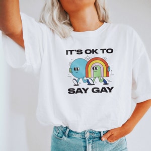 May include: White t-shirt with a graphic of two cartoon characters walking. The characters are a blue circle with a face and a rainbow. The text on the shirt reads "It's OK to Say Gay."