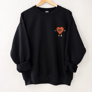 K&ouml;nnte beinhalten: Schwarzer Pullover mit einem Cartoon-Herz-Design. Das Herz ist orange und hat Augen, einen Mund und Arme. Das Herz zeigt mit beiden H&auml;nden nach oben.
