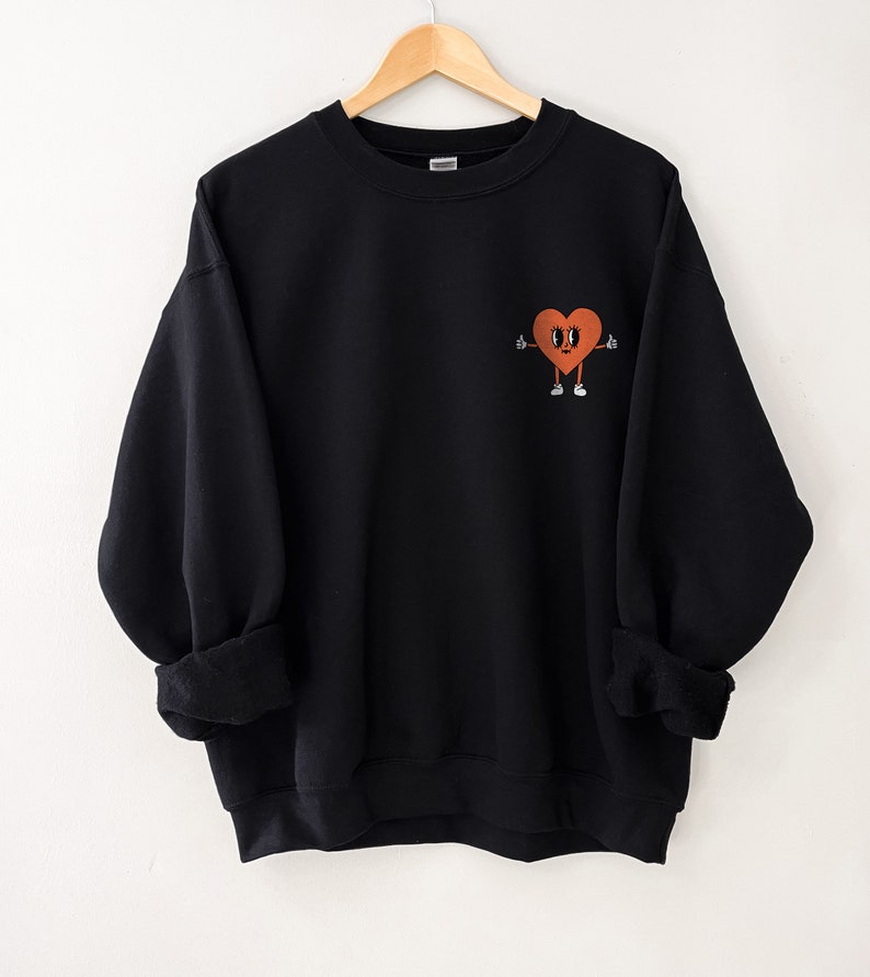 K&ouml;nnte beinhalten: Schwarzer Pullover mit einem Cartoon-Herz-Design. Das Herz ist orange und hat Augen, einen Mund und Arme. Das Herz zeigt mit beiden H&auml;nden nach oben.