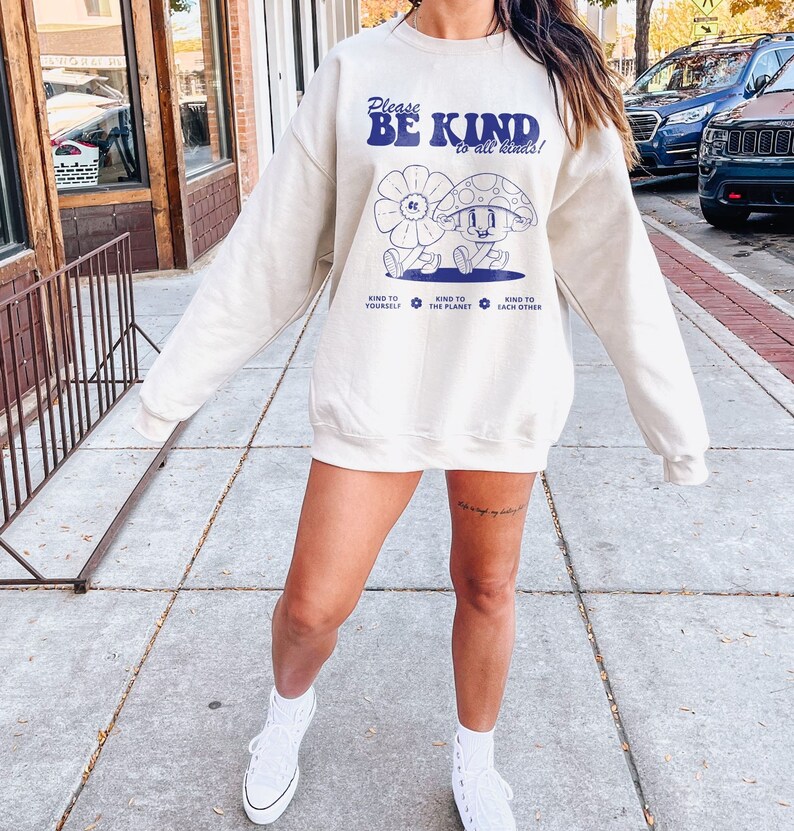 May include: A cream colored sweatshirt with a blue graphic of a mushroom and flower. The text on the sweatshirt reads "Please Be Kind to all kinds!" and "Kind to Yourself, Kind to the Planet, Kind to Each Other".