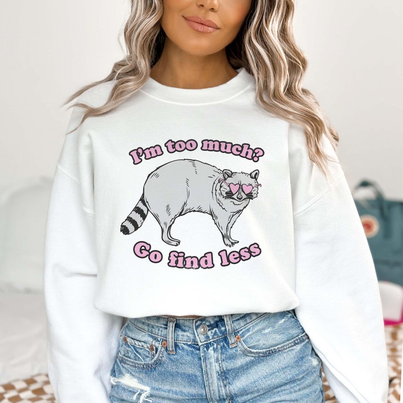 Pu&ograve; includere: Felpa bianca con una grafica di un procione che indossa occhiali da sole a forma di cuore e il testo "I'm too much? Go find less."