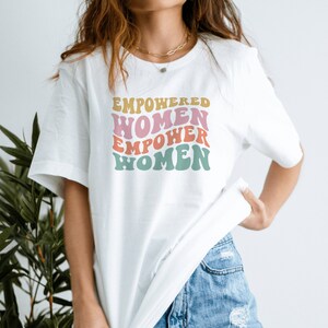May include: White t-shirt with a graphic print that says "EMPOWERED WOMEN EMPOWER WOMEN" in a retro font with pink, yellow, and blue colors.